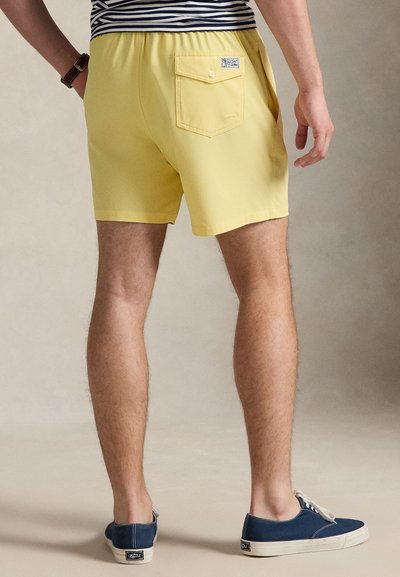 Κίτρινα μαγιό σορτς με ελαστική μέση, πίσω τσέπη και λεία υφή, συνδυασμένα με ναυτικό μπλε καμβά sneakers.