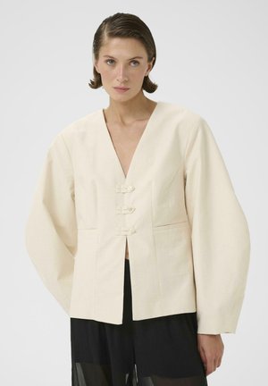 Kvinde med kort brunt hår iført en cremefarvet oversized jakke med tre knapper foran og sorte bukser med vidde, stående mod en ensfarvet baggrund.