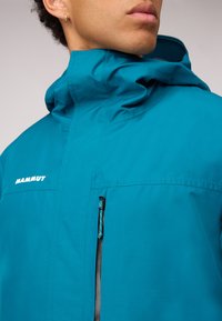 Tealfarbene Mammut-Jacke mit hohem Kragen, Front-Reißverschlusstasche und Logo-Detail. Glatte Textur und modernes Design für den Außeneinsatz.