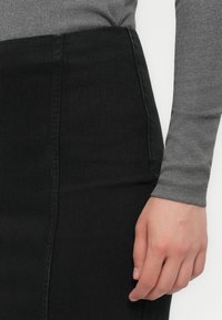 Mână odihnindu-se pe lângă corp, purtând un top gri cu dungi lungi și o fustă din denim neagră cu talie înaltă.
