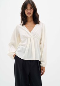 Blusa di colore crema in tessuto leggero con scollatura a V profonda, dettagli plissettati sul davanti e maniche lunghe con polsini elasticizzati, abbinata a pantaloni neri.