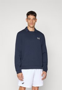 Temno modra polo majica s dolgimi rokavi in ovratnikom, z logotipom EA7 na levi strani prsnega dela ter sproščenim krojem. Spodaj bele športne kratke hlače.
