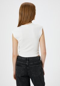 Hvid ribbet ærmeløs top med rund halsudskæring, parret med højtaljede mørkegrå jeans med baglommer og en let slidt tekstur.