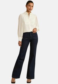 Blusa bianca con dettagli in pizzo sul petto, abbinata a jeans svasati blu scuro. Modella che indossa sandali con zeppa color beige.