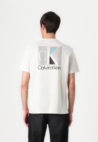Bílý bavlněný tričko s perforovanou grafikou v šedé, modré a černé barvě, se textem "Calvin Klein" umístěným uprostřed zad.