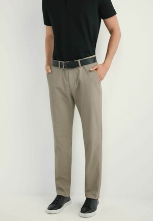 Mężczyzna w beżowych spodniach, czarnym pasku, czarnej koszulce i czarnych trampkach z białą podeszwą, stojący z rękami w kieszeniach przy białej ścianie.