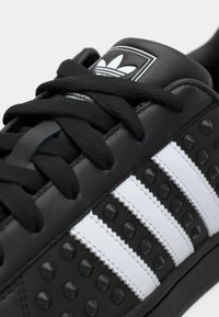 Baskets en cuir noir avec des accents texturés, trois bandes blanches et une languette rembourrée avec logo. Lacets noirs et détails cloutés.