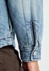 Manga de chaqueta de mezclilla azul claro con cierre de botón y costuras en contraste, con un acabado texturizado y detalle de abertura lateral.