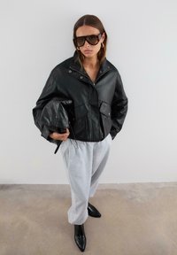 Žena nosi oversize crnu kožnu jaknu, svijetlosive trenirke, crne štikle na špic, velike sunčane naočale, drži crnu kožnu torbu.