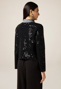 Schwarze, mit Pailletten verzierte kurze Jacke mit einem glatten grauen Kragen, kombiniert mit schwarzen Hosen. Verfügt über lange Ärmel und ein minimalistisches Design.