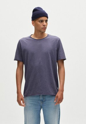 Lila t-shirt med rund hals i mjukt tyg, korta ärmar och avslappnad passform, matchad med ljusblå jeans och en marinblå mössa.