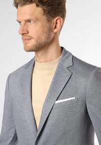 Herrenblazer in Grau mit Muster, ausgestattet mit einem Reverskragen, einem weißen Einstecktuch und einem weichen beigen T-Shirt darunter.