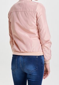 Kvinna iklädd en ljusrosa bomberjacka och mörkblå denimjeans, sedd bakifrån mot en enfärgad bakgrund.