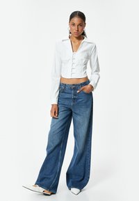 Camicetta bianca corta con chiusura a bottoni, abbinata a jeans blue in denim a gamba larga. La camicia presenta un colletto e maniche lunghe.
