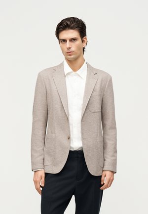 Homme portant un blazer beige texturé sur une chemise blanche boutonnée et un pantalon noir, debout devant un fond clair uni.