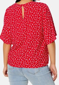 Blouse rouge à motifs floraux avec des manches courtes et flottantes, dotée d'un détail en clé au dos, avec de petits motifs de fleurs blanches et bleues sur un tissu doux.