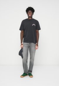 Sort bomuld t-shirt med korte ærmer og trykt detalje, parret med grå jeans og grønne sneakers med brune accenter. Kasket i hånden.