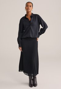 Donna che indossa una blusa nera con bottoni, una gonna midi plissettata nera e stivaletti neri in pelle, in piedi con una mano sulla vita.