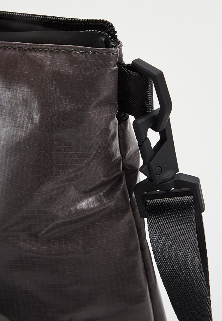 Schwarze Nylon-Tasche mit Reißverschluss und verstellbarem Gurt, die an einem Plastikdrehhaken vor einem neutralen Hintergrund befestigt ist.