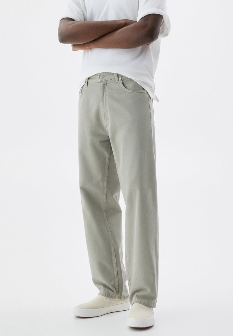 Personne portant un jean droit gris clair, des baskets blanches sans lacets et une chemise blanche à manches courtes, bras croisés.