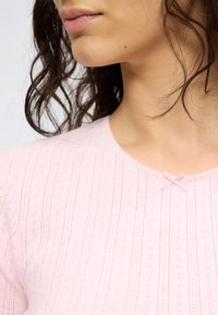 Γυναίκα με κυματιστά σκούρα μαλλιά φοράει ένα ανοιχτό ροζ πλεκτό τοπ με υφή και μικρό φιόγκο στο λαιμό.