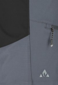 Nahaufnahme von grauem und schwarzem Stoff mit einem Reißverschluss und einem dreieckigen Logo, das nahe am unteren Rand eingestickt ist.