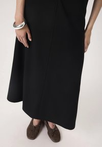 Czarna sukienka do kolan z gładkiego materiału o lekko A-liniowym kształcie. Modelka ma na sobie brązowe zamszowe buty i srebrną bransoletkę na nadgarstku.
