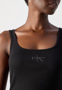 Gros plan sur une personne portant un débardeur noir à côtes avec le logo Calvin Klein, une boucle d'oreille, et des lèvres visibles sur fond blanc.
