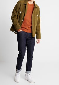 Person i olivgrön knappad jacka, orange stickad tröja, mörka uppvikta jeans och vita sneakers som står mot en enkel bakgrund.