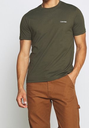 Mężczyzna w oliwkowozielonej koszulce Calvin Klein i brązowych spodniach, stojący z jedną ręką w kieszeni na jednolitym tle.