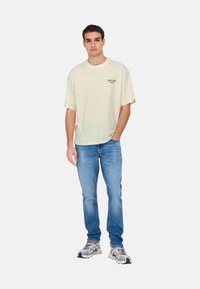 Lys beige oversized t-shirt med broderet brunt logo, parret med blå jeans og grå sneakers med sorte accenter og strukturerede overflader.