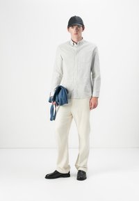 Joven vistiendo una camisa de botones de rayas claras, pantalones crema, gorra negra y zapatos negros, sosteniendo una chaqueta azul doblada en su mano izquierda.