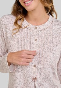 Mujer sonriendo, abrochándose una blusa ligera de color rosa con un patrón y pliegues en pincelada, con un pequeño detalle de lazo en el dobladillo.