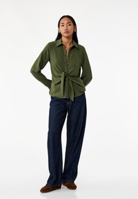 Olivgrön skjorta med knappar och knytdetalj fram, långa ärmar och strukturerad tyg, matchas med lösa mörkblå jeans och bruna skor.