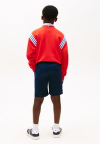 Chlapec stojaci chrbtom, oblečený v červenej mikine s dlhými rukávmi s bielymi a modrými pruhmi, tmavomodrých šortkách, bielych ponožkách a teniskách.