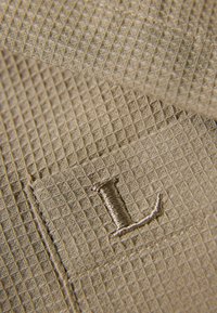 Gros plan sur un tissu beige à texture gaufrée avec la lettre majuscule « L » brodée et des coutures visibles le long des bords.