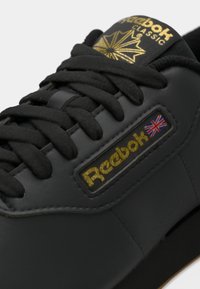 Sapatilha de couro preto com atacadores pretos, um logotipo dourado "Reebok Classic" e um detalhe da bandeira britânica. Apresenta uma superfície texturizada e lingueta almofadada.