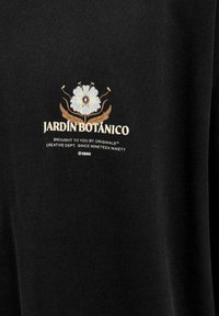 Must kaelus T-särk, millel on lilleline graafika tekstiga "JARDÍN BOTÁNICO" beeži ja helesinise värviga. Pehme tekstuur, klassikaline lõige.