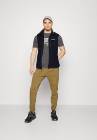 Schwarze Fleece-Weste über einem grauen Grafik-T-Shirt, kombiniert mit beigen Jogginghosen und schwarzen Sportschuhen. Der Hintergrund ist schlicht weiß.