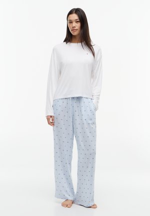 Femme debout pieds nus portant une chemise blanche unie à manches longues et un pantalon de pyjama ample bleu clair avec un petit motif carré.