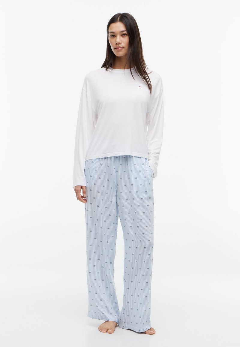 Femme debout pieds nus portant une chemise blanche unie à manches longues et un pantalon de pyjama ample bleu clair avec un petit motif carré.