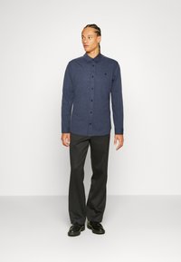 Long-sleeve navy button-up shirt with a textured finish, and dark trousers. Shirt features a collar and logo accent on the chest.