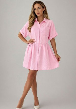 Femme portant une robe rose à manches courtes, boutonnée devant, avec col et poche, posant avec une main sur la taille et des talons blancs.