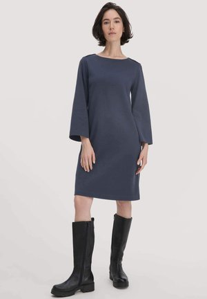 Vrouw staat in een knielange marineblauwe jurk met lange mouwen en zwartekniehoge laarzen tegen een effen achtergrond.