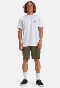 T-shirt bianco in cotone a maniche corte e colletto rotondo, con un logo circolare teal sul lato sinistro del petto; abbinato a pantaloni corti verde oliva.