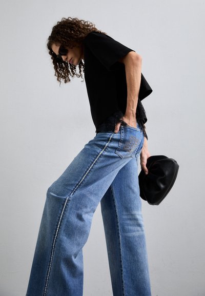 Juodas trumpas viršus su nėrinių apdaila, derinamas su plačiomis šviesiai mėlynomis džinsomis, pasižyminčiomis kontrastingomis siūlėmis ir dekoratyvine užpakaline kišenėle. Laiko juodą rankinę.