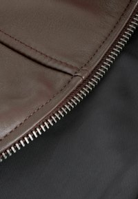 Barna bőr pénztárca látható varrással és ezüst cipzárral, texturált fekete szövet háttér előtt.