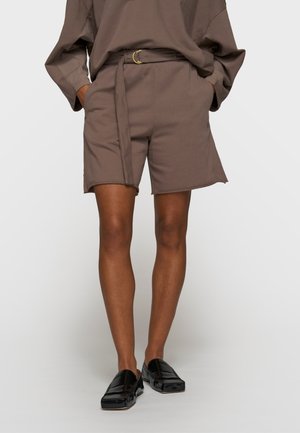 Osoba ubrana w brązowe szorty z paskiem i bocznymi rozcięciami oraz dopasowaną bluzkę z długim rękawem, zestawione z czarnymi wsuwanymi mokasynami.