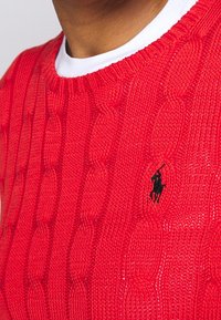 Röd kabelstickad tröja med rund hals, texturerat mönster och en svart broderad logotyp på vänster bröst.