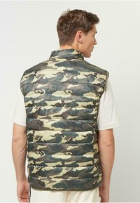 Tarnfarbenes Pufferwesten in Grün- und Beigetönen, aus glänzendem, leichtem Material gefertigt. Verfügt über einen hohen Kragen und ein ärmelloses Design.